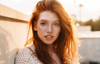 A young woman with long red hair and freckles looks directly at the camera, standing outdoors in warm sunlight next to a concrete wall.