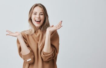 Pretty beautiful woman with blonde long hair looking at camera having excited and happy facial expression, clapping with her hands against blank studio wall, expressing her excitement with present
